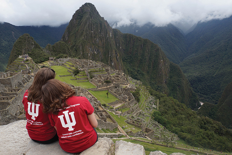 Two female students wearing IU t-shirts overlook mountains and stone structures in another country.