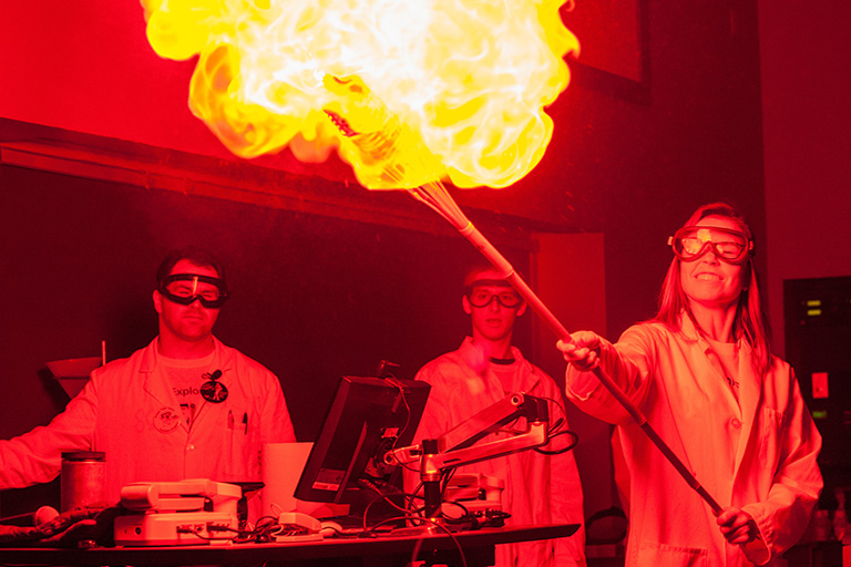 In a red lit room, an enormous flame explodes at the end of a stick held by a faculty member. Two students observe, wearing protective eyewear. 
